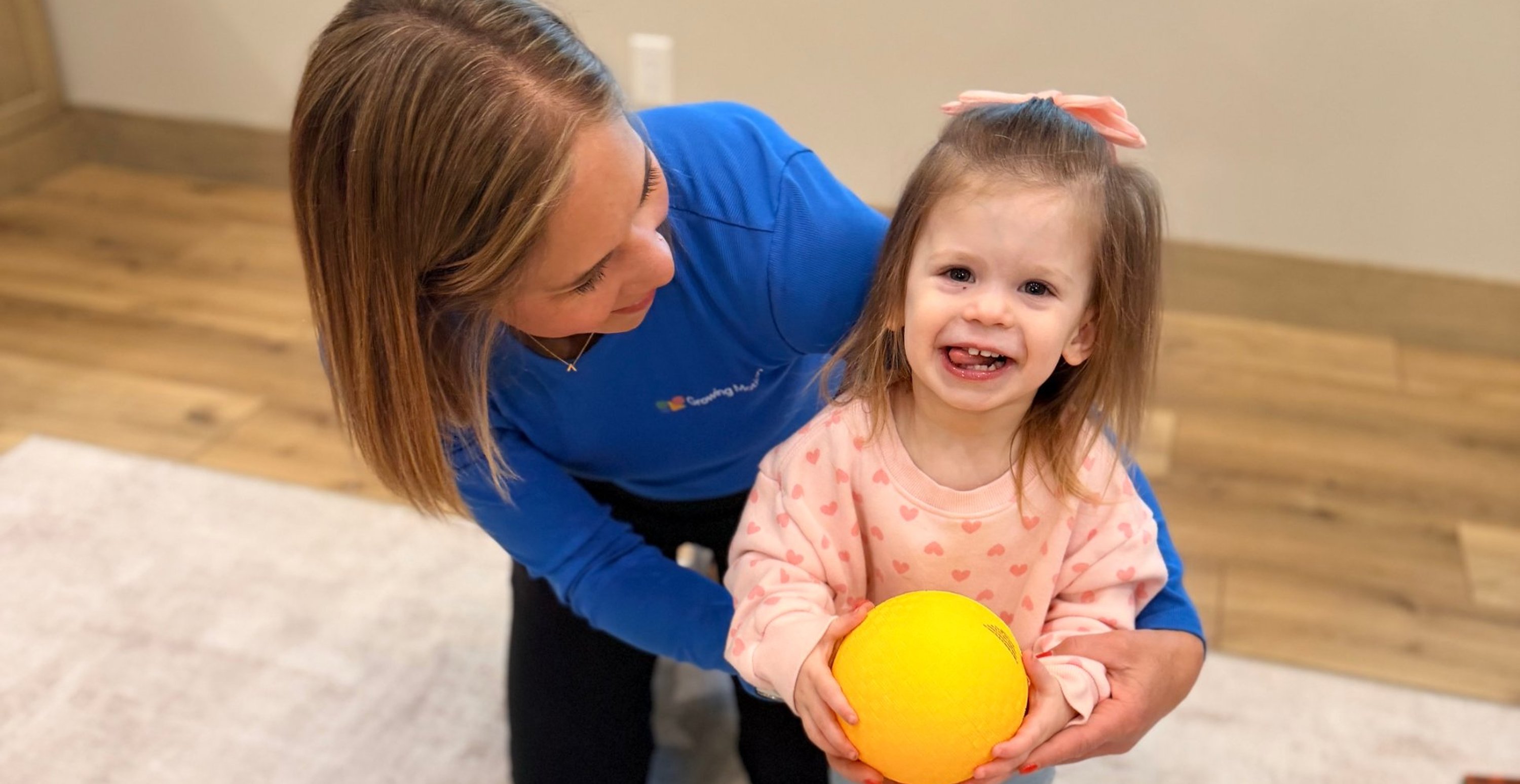 Growing Motion therapist helping a smiling toddler hold a yellow ball during an in-home therapy session